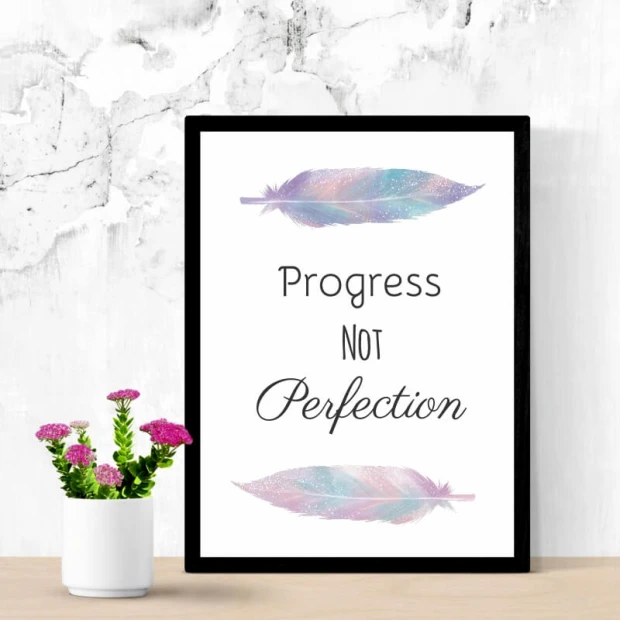 Plakat w czarnej ramie z motywacyjnym napisem Progress not perfection oraz grafiką kolorowych piór