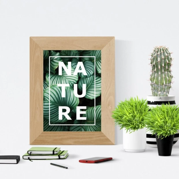 Plakat z motywem zielonych liści i białym napisem NATURE, zaprezentowany w jasnej, drewnianej ramie