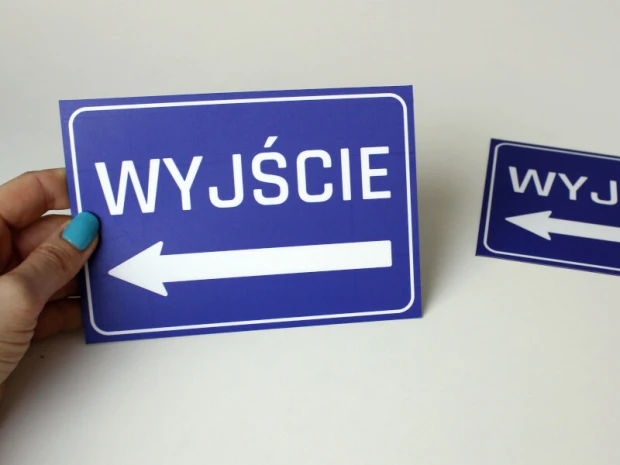 Niebieska naklejka BHP z napisem WYJŚCIE i strzałką w lewo, prezentowana na dłoni jako gotowy produkt