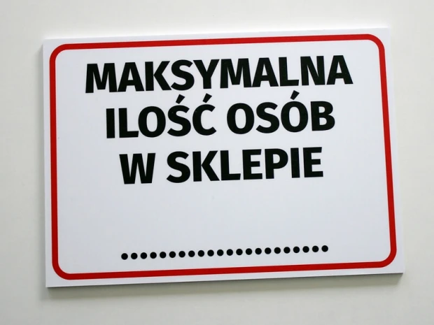 Biała tabliczka BHP z czerwonym obramowaniem i napisem Maksymalna ilość osób w sklepie zamontowana na ścianie