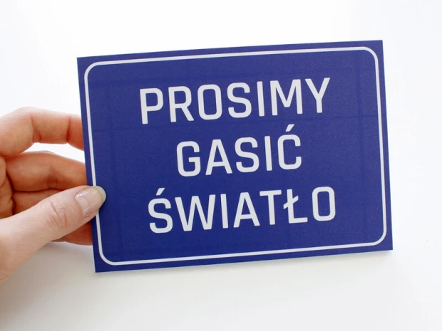 Niebieska tabliczka BHP Prosimy gasić światło trzymana w dłoni, co obrazuje jej rzeczywisty rozmiar