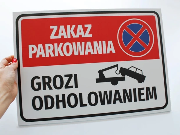 Tabliczka Zakaz parkowania grozi odholowaniem trzymana w dłoni, co obrazuje jej rzeczywisty rozmiar