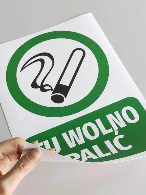 Zielona naklejka BHP TU WOLNO PALIĆ z piktogramem papierosa podczas odklejania od papieru podkładowego