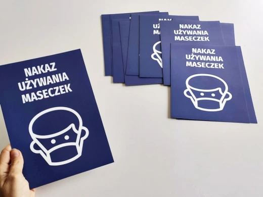 Niebieska naklejka BHP do biur i sklepów z białym piktogramem i napisem Nakaz używania maseczek, trzymana w dłoni