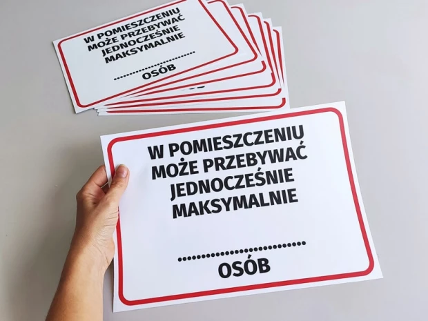 Naklejka BHP z limitem osób w czerwonej ramce, trzymana w dłoni na tle stosu gotowych naklejek