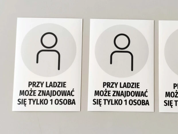 Trzy naklejki BHP z szarym piktogramem postaci i napisem informacyjnym naklejone obok siebie na gładkiej powierzchni