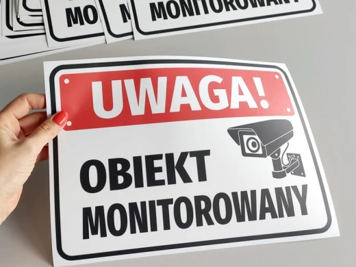 Naklejka BHP Uwaga obiekt monitorowany z czerwoną belką i kamerą, pokazana jako elastyczny arkusz w dłoni