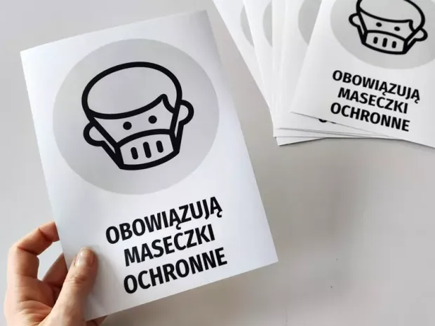 Szaro-czarna naklejka BHP do firmy z napisem Obowiązują maseczki ochronne trzymana w dłoni