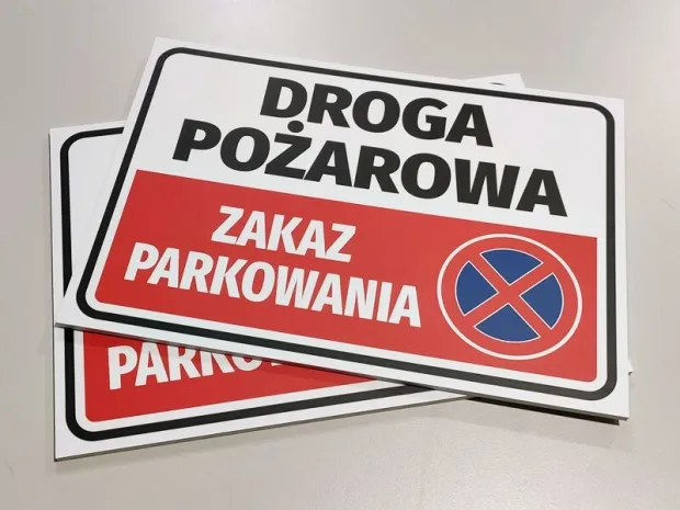 Trzy tabliczki Droga pożarowa zakaz parkowania ze znakiem zakazu postoju, ułożone w stos na płaskiej powierzchni