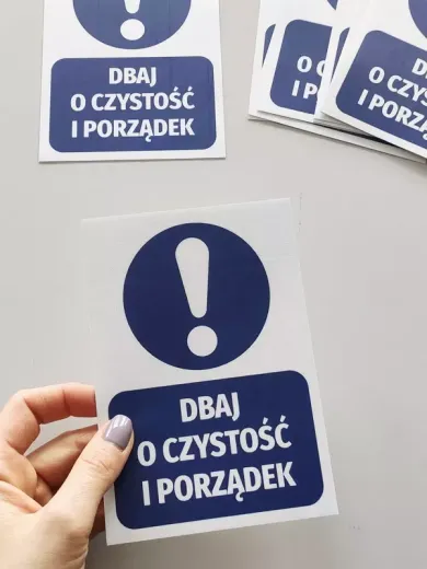 Niebieska naklejka BHP nakazu dbania o czystość z wykrzyknikiem, trzymana w ręku