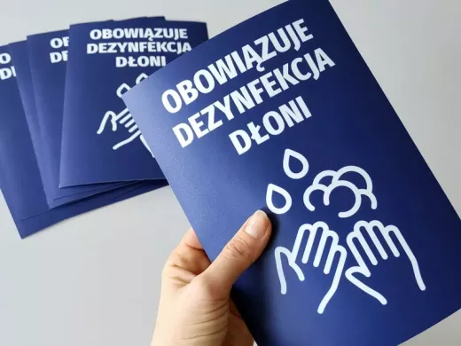 Niebieskie naklejki BHP z nakazem dezynfekcji dłoni i piktogramem rąk do biura, trzymane w dłoni