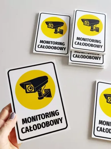 Tabliczka z napisem MONITORING CAŁODOBOWY i ikoną kamery, prezentowana w dłoni oraz w ułożonym stosie