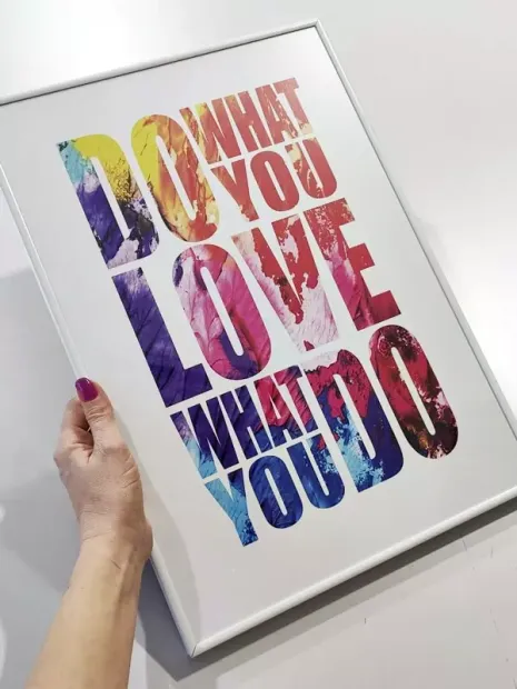 Plakat w białej ramie z kolorowym napisem DO WHAT YOU LOVE WHAT YOU DO trzymany w dłoni na jasnym tle