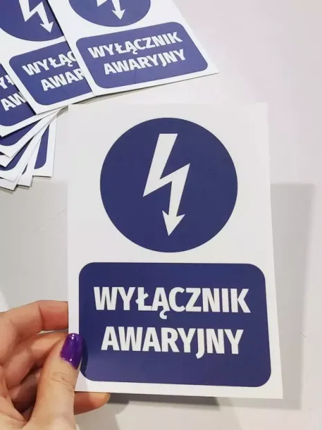 Niebieska naklejka BHP Wyłącznik awaryjny z błyskawicą do instalacji technicznych, trzymana w dłoni