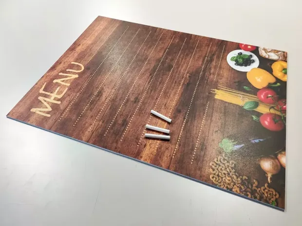 Tablica kredowa z nadrukiem menu, grafiką warzyw i trzema kawałkami kredy w ujęciu pokazującym grubość płyty