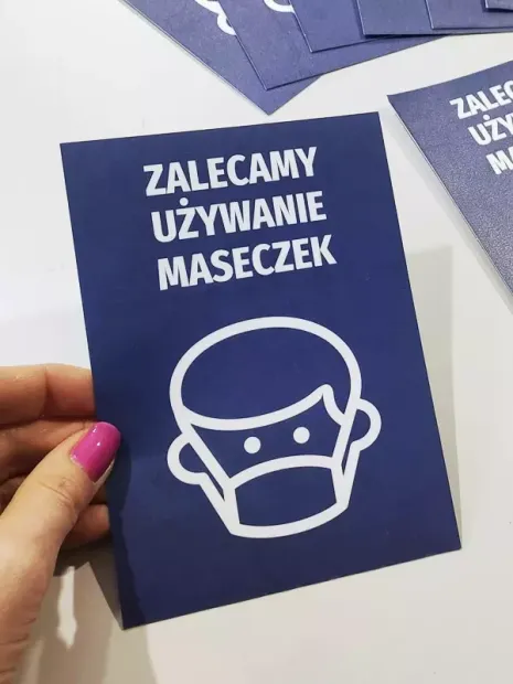 Niebieska naklejka BHP z napisem Zalecamy używanie maseczek, pokazana w dłoni jako fizyczny produkt