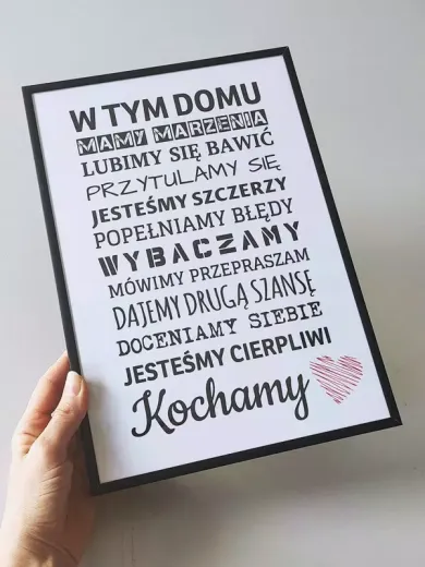 Plakat w czarnej ramie z napisem W tym domu i różowym sercem trzymany w dłoni, pokazujący fizyczny produkt