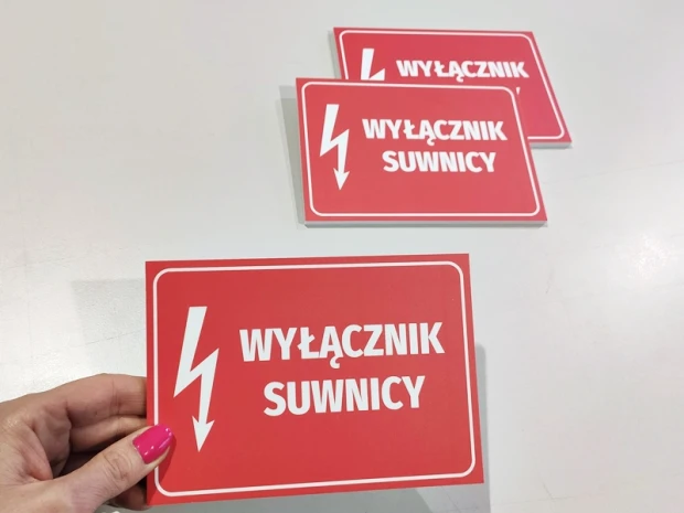 Trzy czerwone tabliczki BHP z napisem Wyłącznik suwnicy i piktogramem błyskawicy, trzymane w dłoni