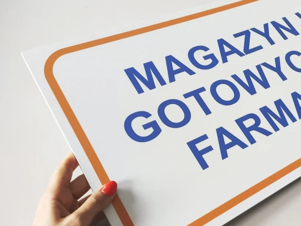 Tabliczka z napisem MAGAZYN WYROBÓW GOTOWYCH PRODUKCJI FARMACEUTYCZNEJ trzymana w dłoni, pokazująca grubość płyty
