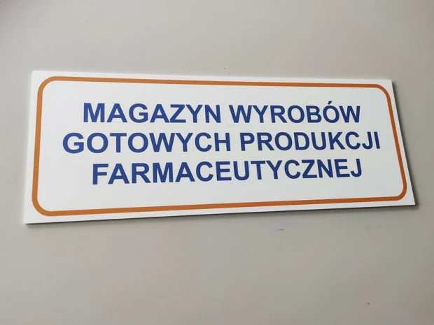 Prostokątna tabliczka z napisem MAGAZYN WYROBÓW GOTOWYCH PRODUKCJI FARMACEUTYCZNEJ zamontowana na ścianie