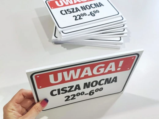 Tabliczka informacyjna UWAGA! CISZA NOCNA 22:00-6:00 trzymana w dłoni na tle stosu gotowych produktów