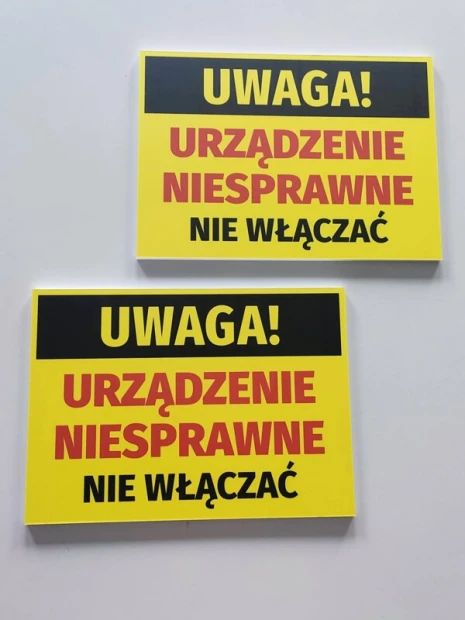 Dwie żółte tabliczki ostrzegawcze BHP z napisem UWAGA! URZĄDZENIE NIESPRAWNE NIE WŁĄCZAĆ na gładkiej powierzchni
