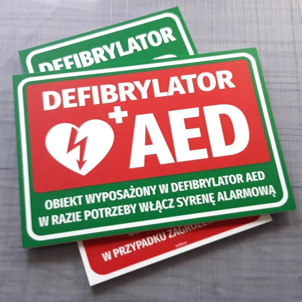 Tabliczka informacyjna Defibrylator AED - Wzór graficzny. Prostokątna tabliczka z czerwonym tłem, białym tekstem i symbolem